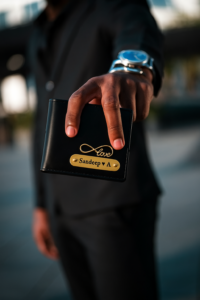 A hand in a dark suit extends a black leather wallet. The wallet has a golden infinity symbol intertwined with the word “love” and a brass nameplate engraved with “Sandeep ♥ A.” Customised Wallet for Men