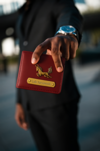 A person in a dark suit holds a red leather wallet featuring a golden horse emblem and a brass nameplate engraved with "Lalit Suryawnshi." The focus is on the wallet while the person’s face is blurred in the background. Customised Wallet for Men